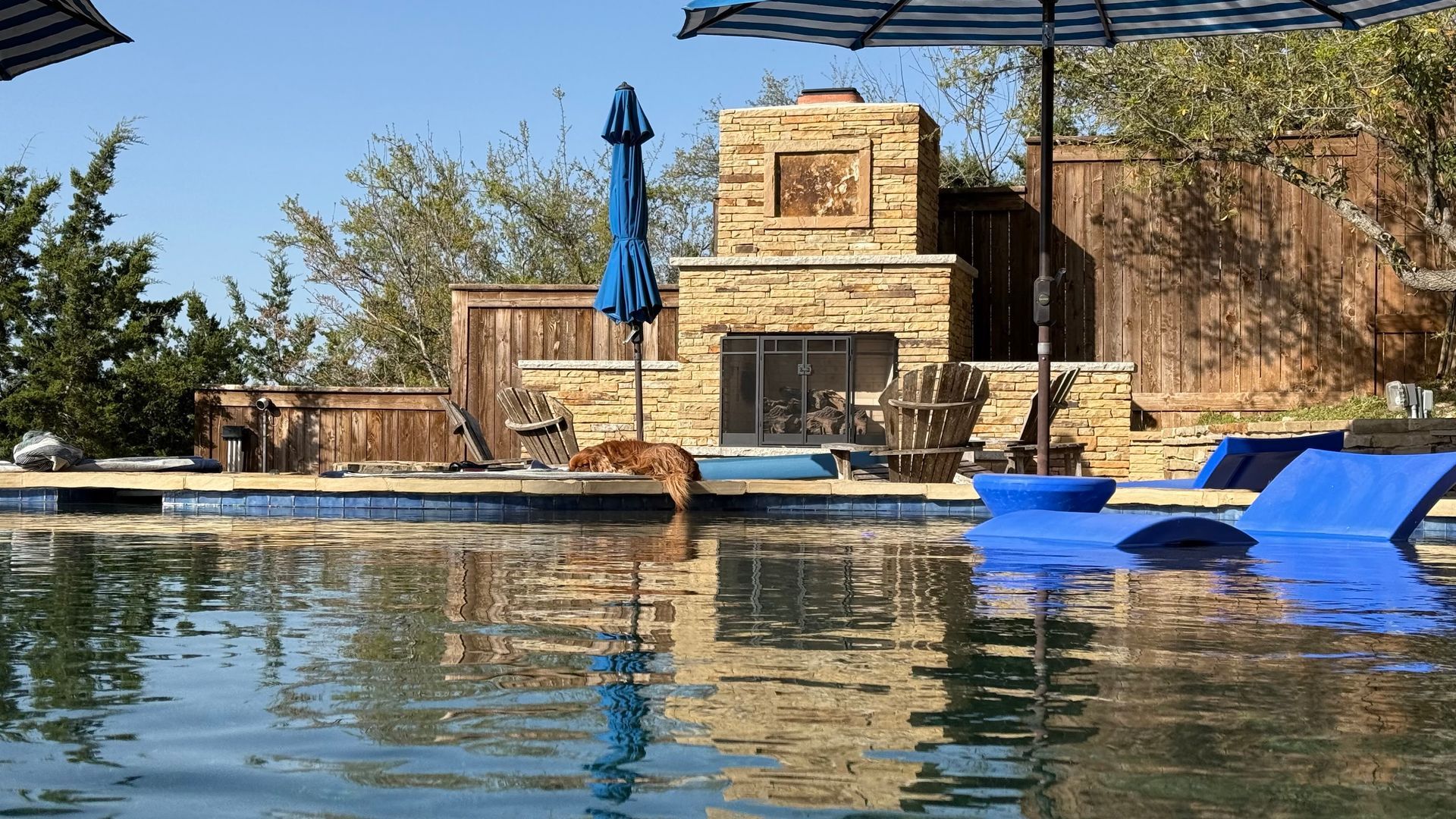 A swimming pool with reflections of a stone fireplace, blue umbrellas, and lounge chairs on a sunny day.