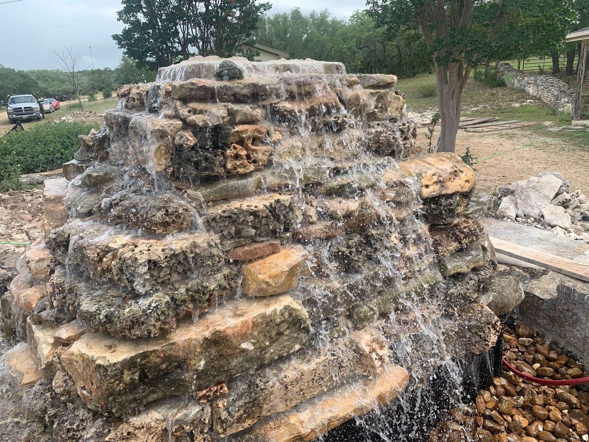 Water cascading down a tiered rock fountain outdoors; a truck and trees are visible in the background.