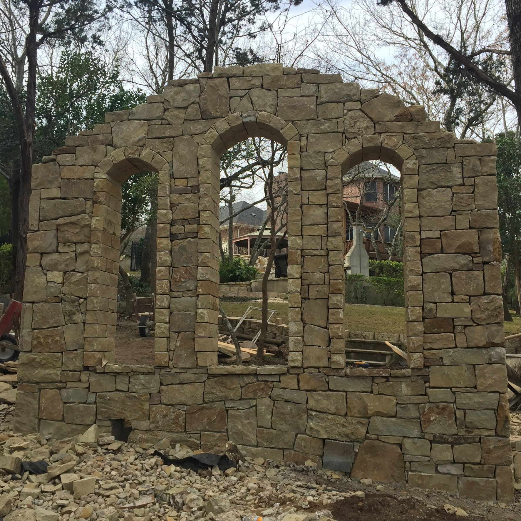 Stone wall with three arched windows; trees and a building are visible behind it.