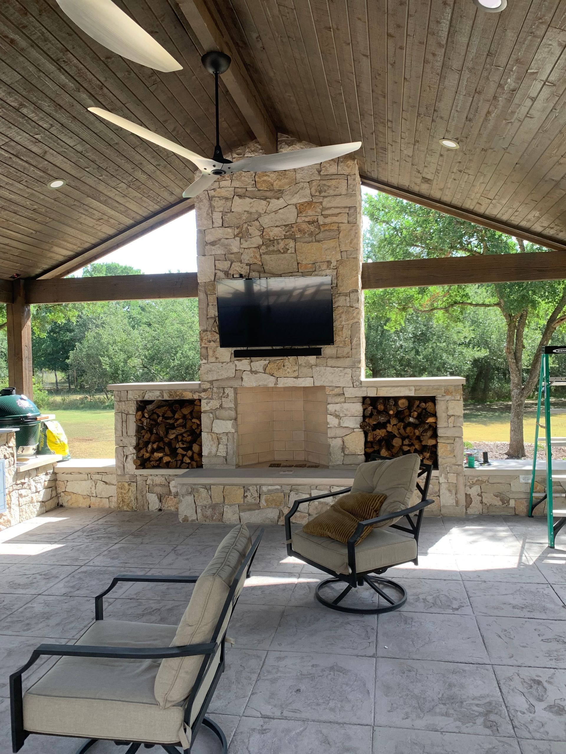 Covered outdoor patio with stone fireplace, TV, seating, and a ceiling fan.