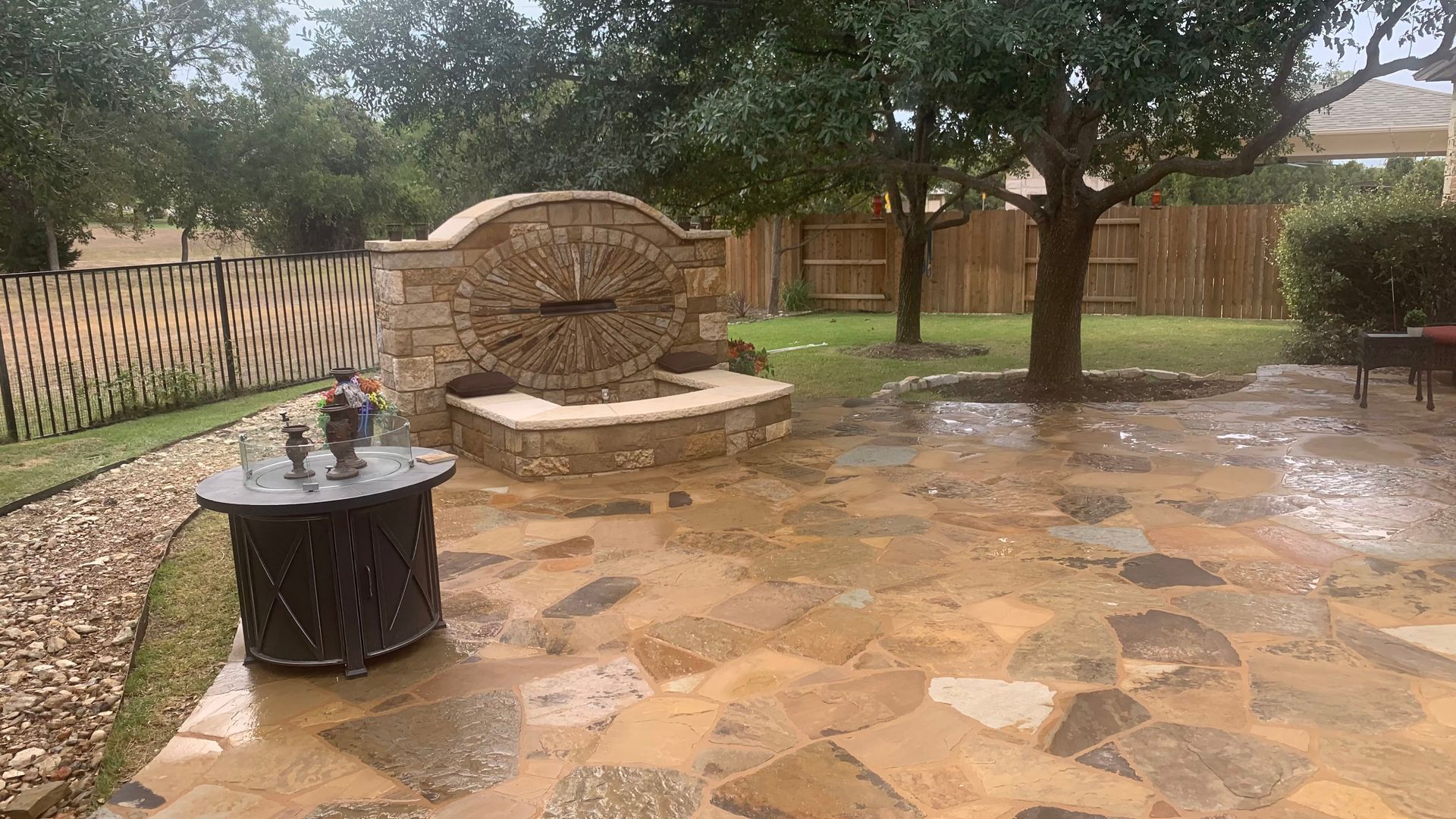 Stone patio with a water fountain and a table. It's raining. Green trees and a wooden fence are visible.