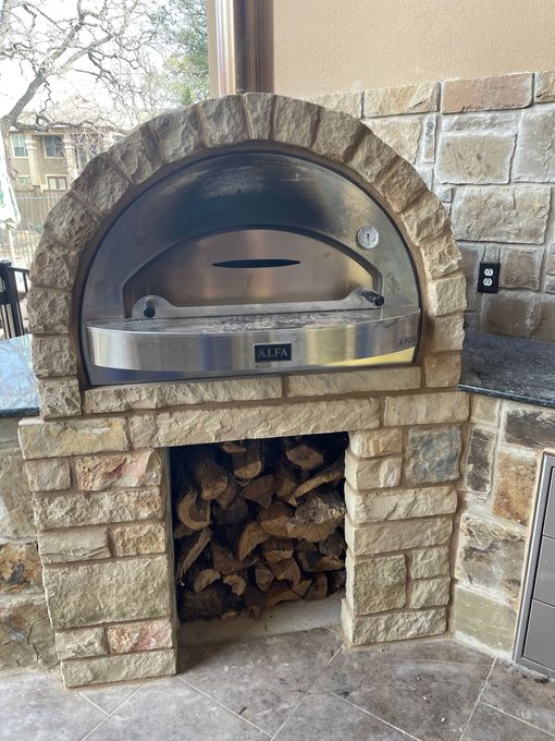 Wood-fired pizza oven built into a stone structure with firewood storage below and a temperature gauge on the oven door.