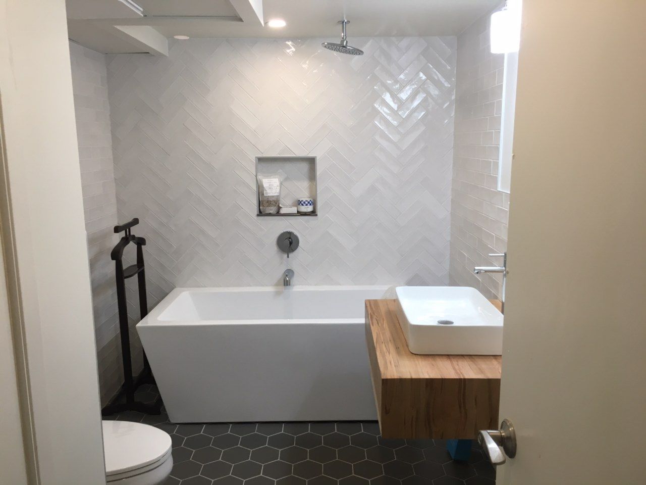 Modern bathroom with white tub, sink, and herringbone tile, featuring a wooden countertop and dark hexagonal floor.
