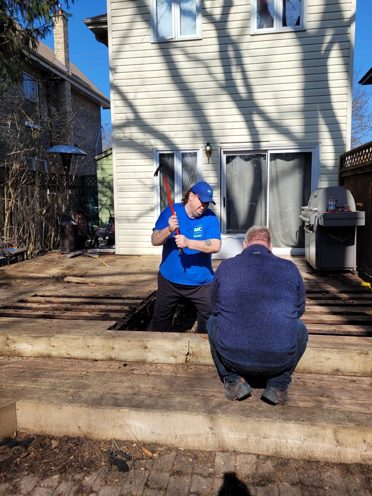 Two men are working on a wooden deck in front of a house.