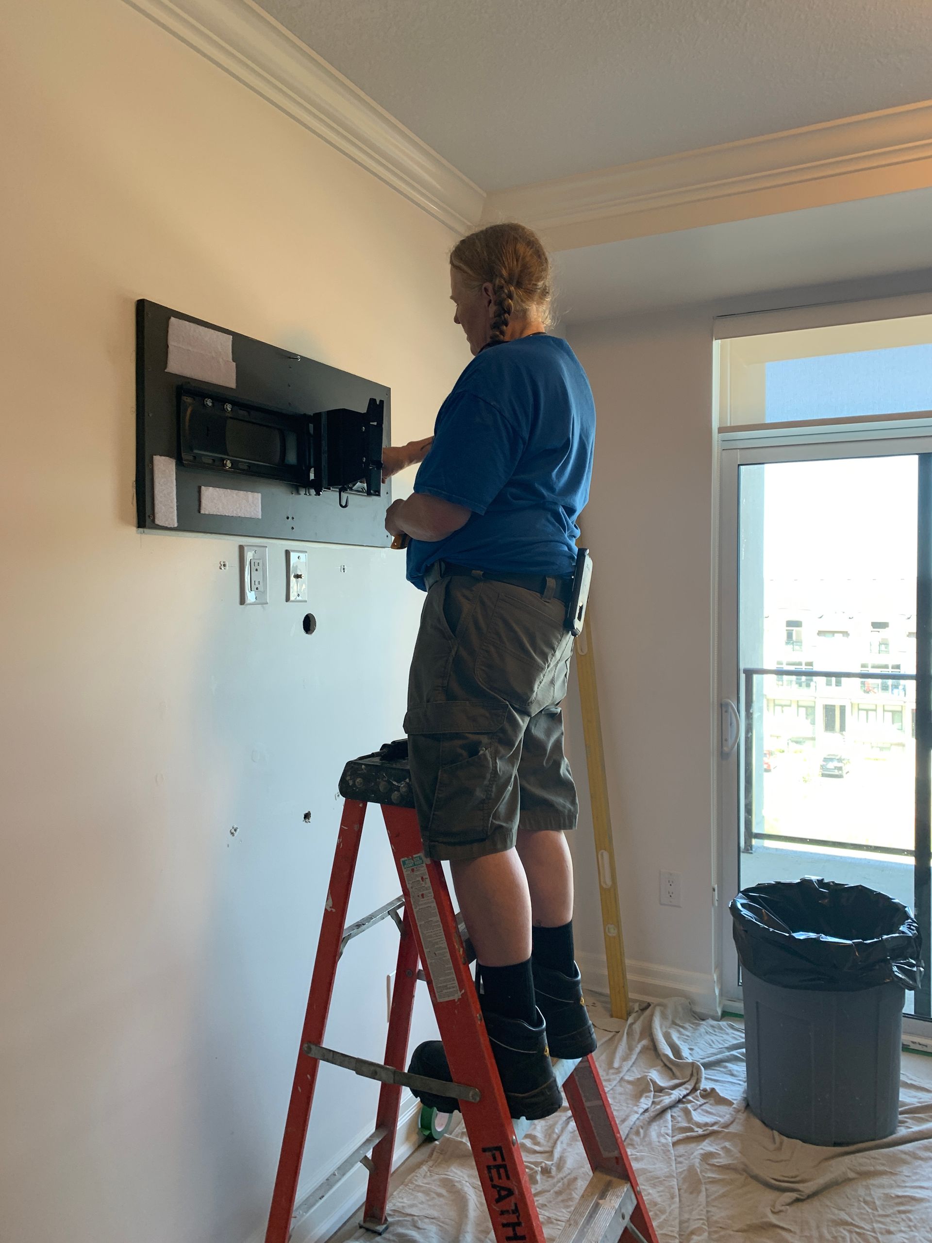 A man is standing on a ladder fixing a television on a wall.