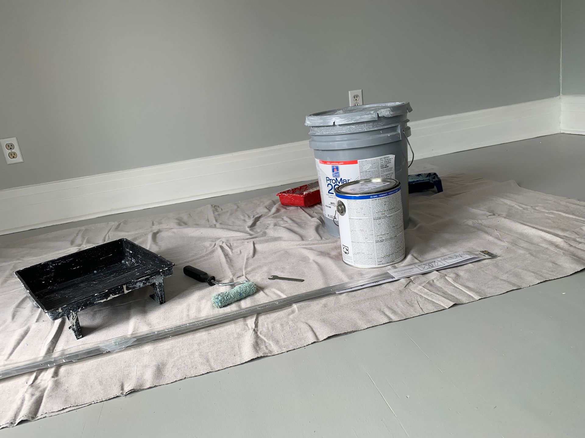 A room with a bucket of paint , a roller , and a tray on the floor.