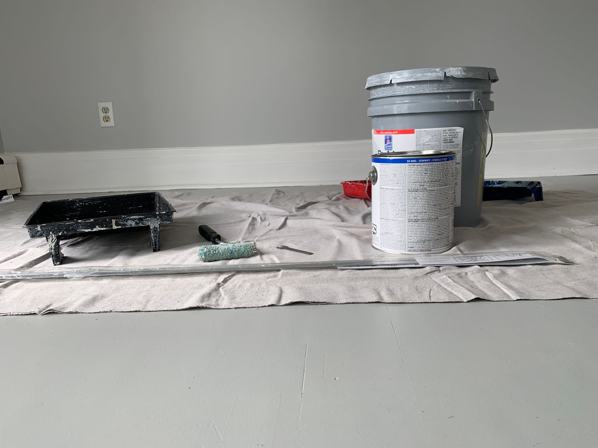 A bucket of paint , a roller , and a brush are on the floor.
