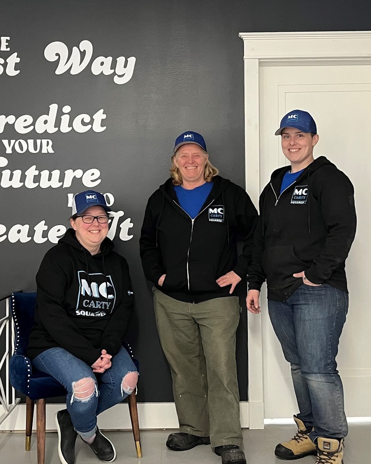 Three people in black hoodies and blue hats stand near a wall with text.