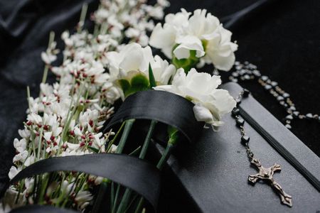 White flowers wrapped in black ribbon, resting beside a dark, rectangular object and a rosary on a black surface.