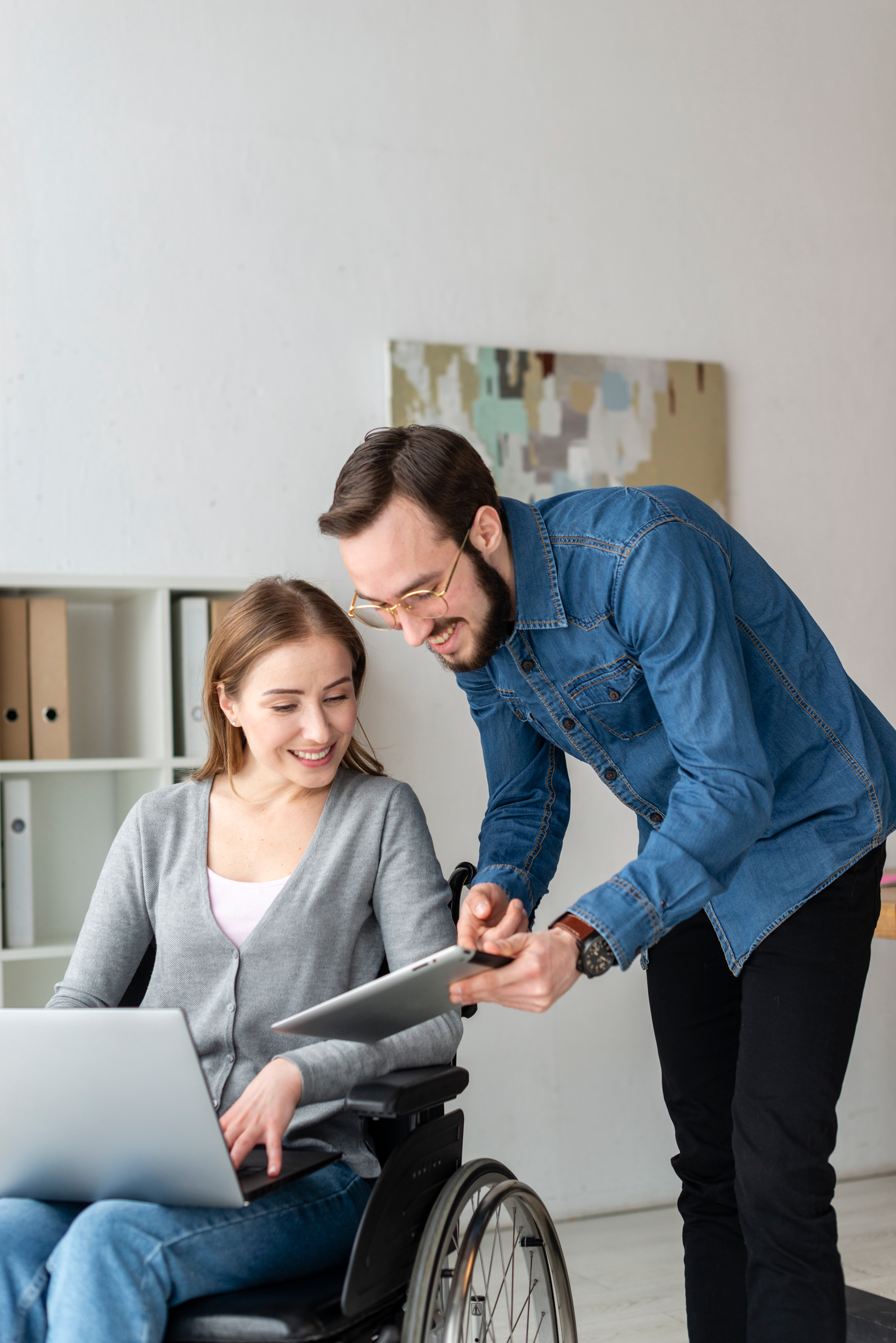 Woman in wheelchair uses laptop while man points to tablet, office setting.