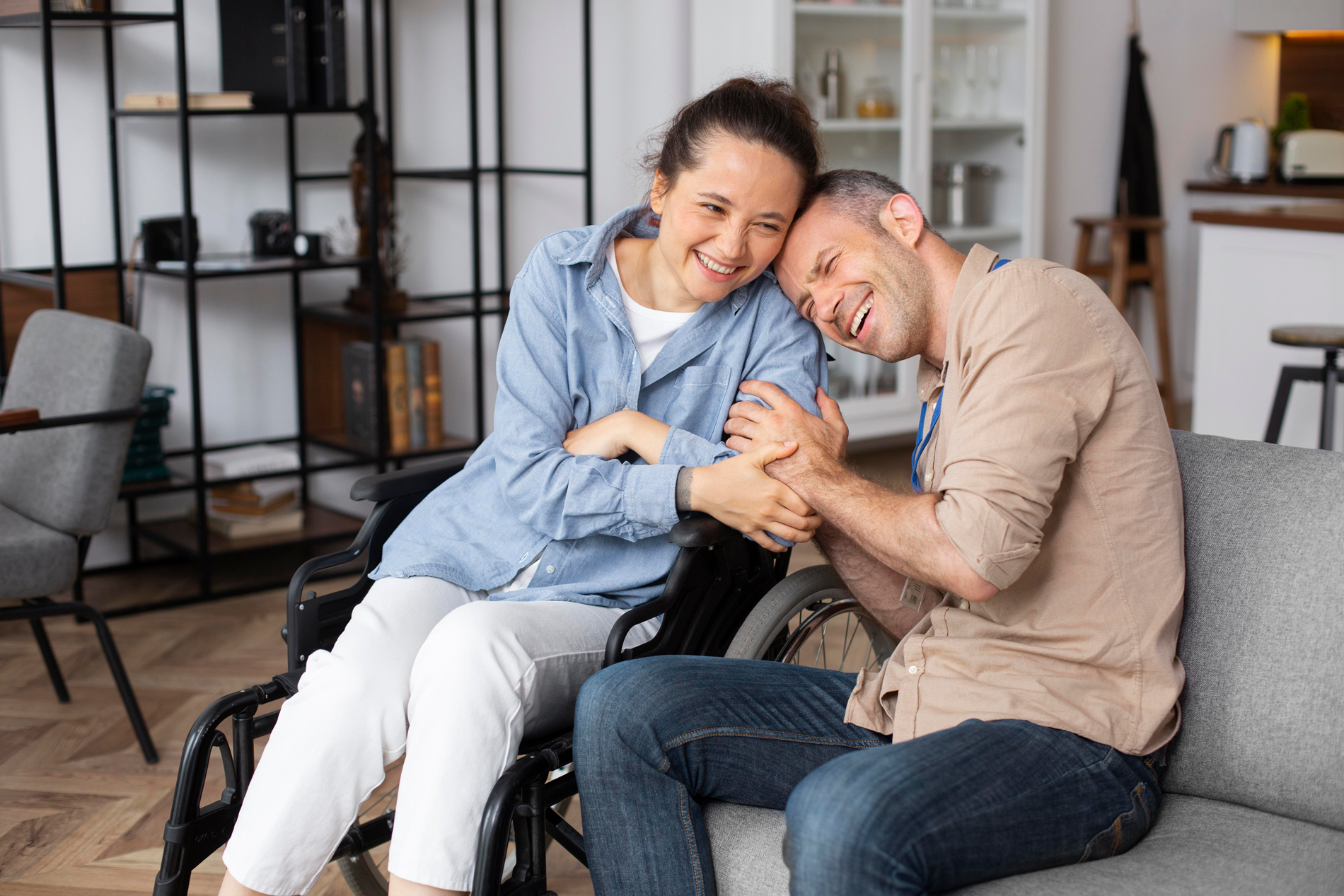 Woman in wheelchair and man on couch holding hands, smiling indoors.