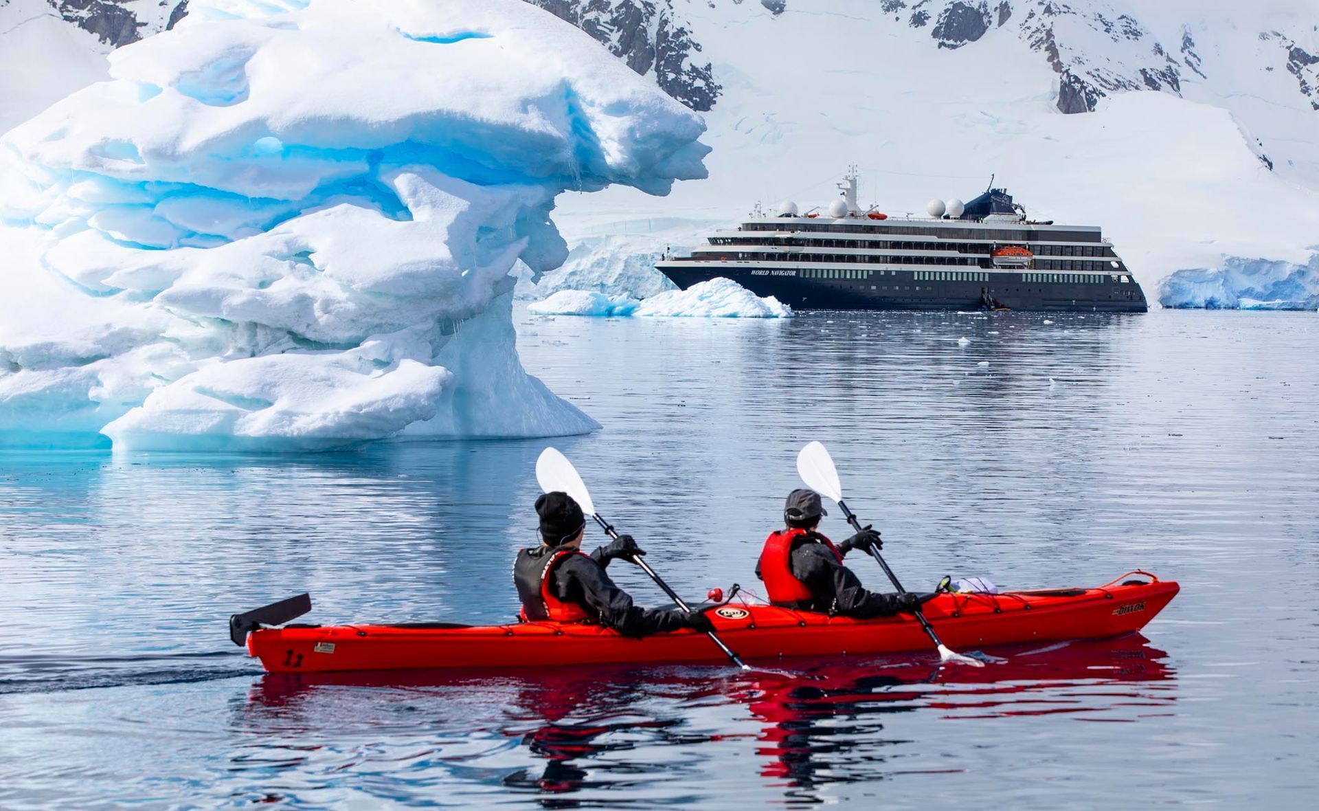 Two people are kayaking in the water with a cruise ship in the background.