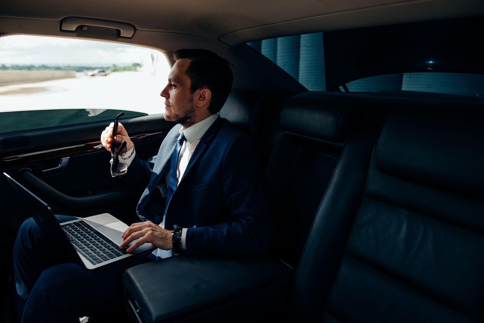 A man in a suit is sitting in the back seat of a car using a laptop computer.