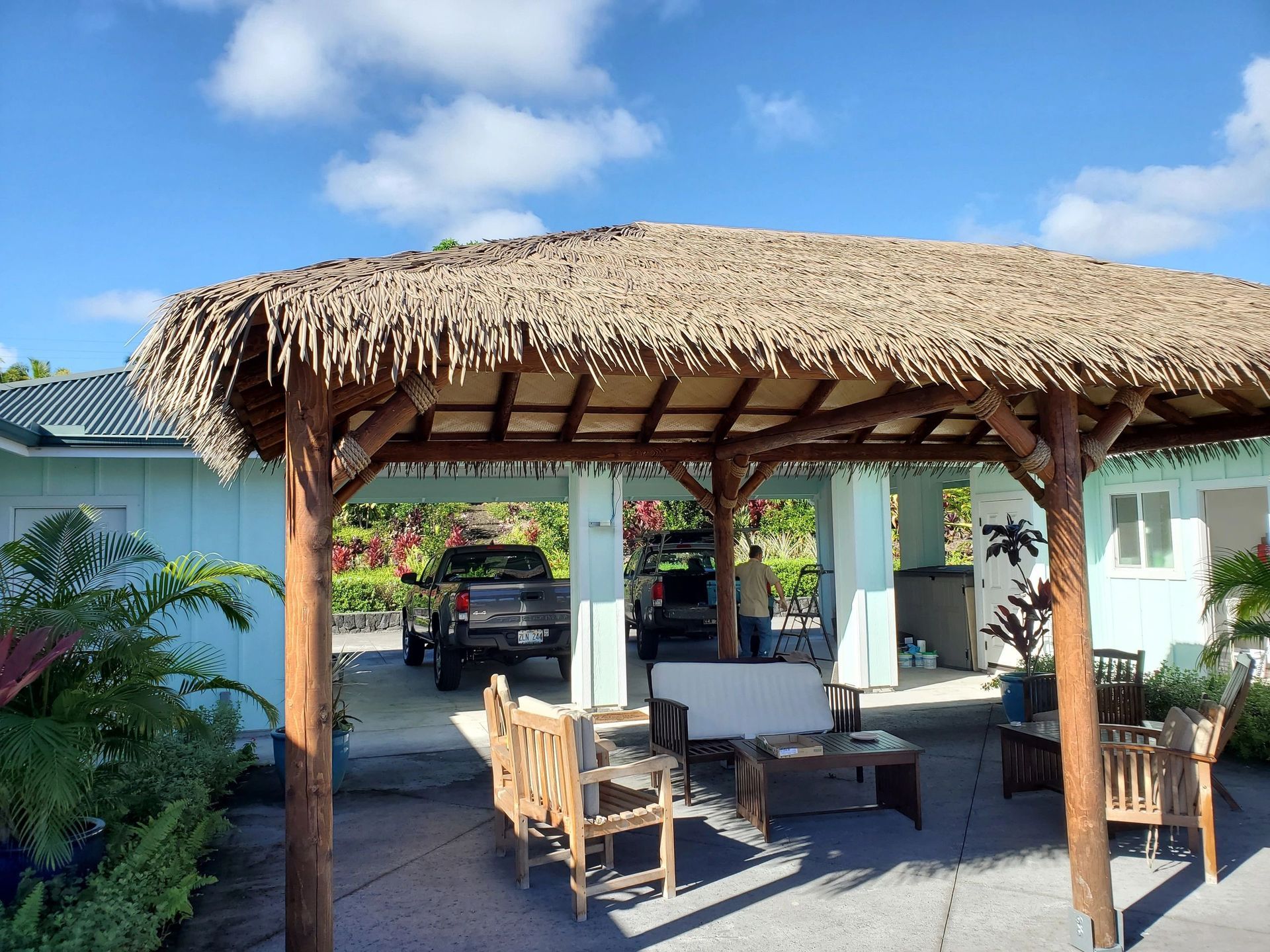 Tiki hut with thatched roof covers outdoor seating area and parked vehicles; blue building in background.