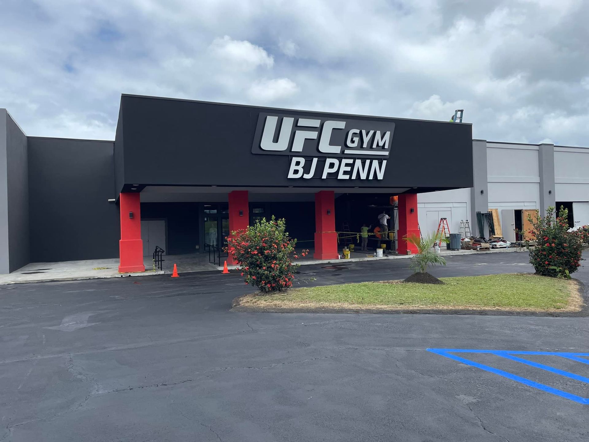 UFC Gym exterior with black signage, red pillars, and BJ Penn's name. Entrance with workers and some landscaping in front.
