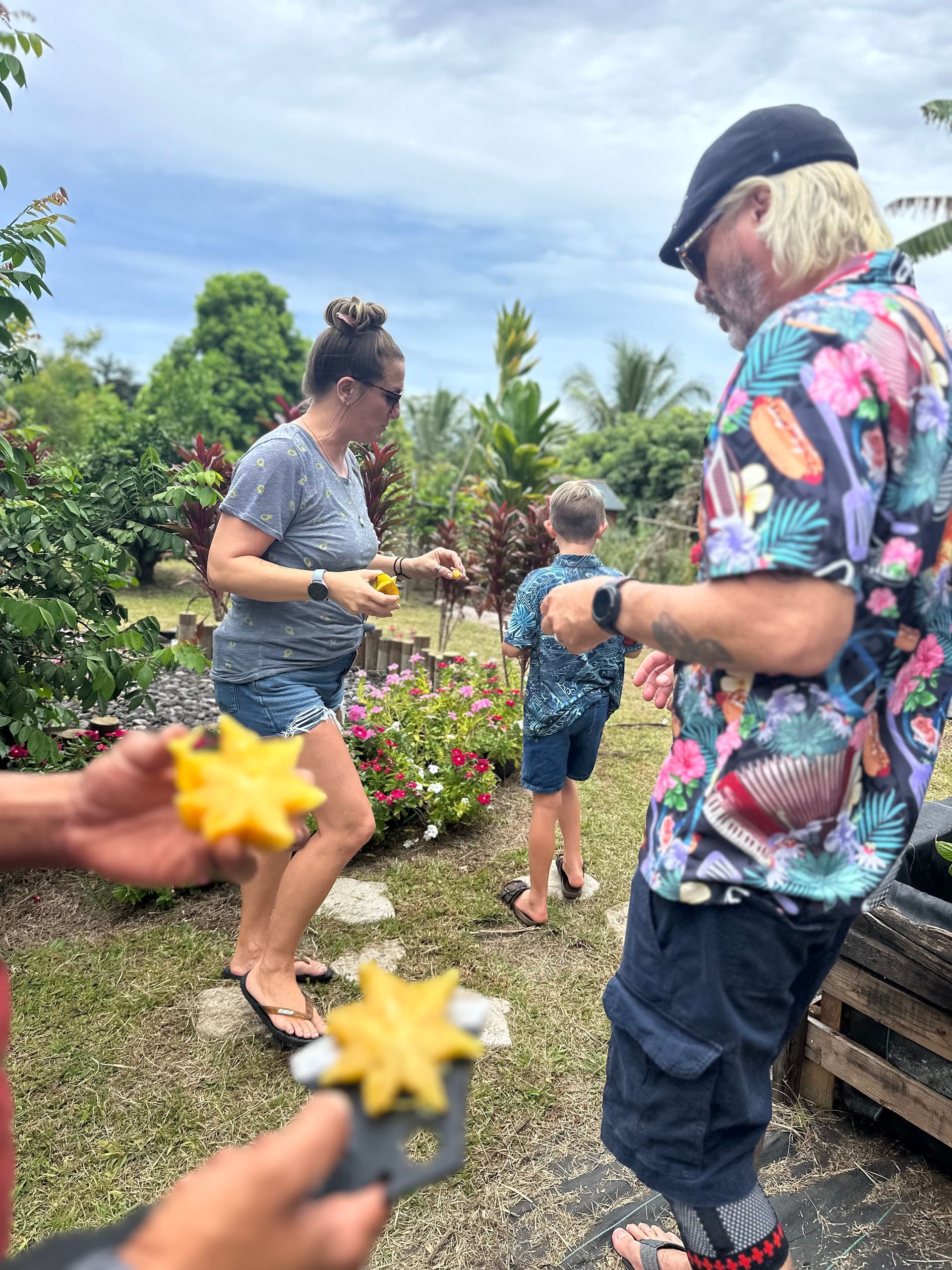 People in a garden, holding starfruit. One person in a floral shirt. Cloudy day with flowers and trees.
