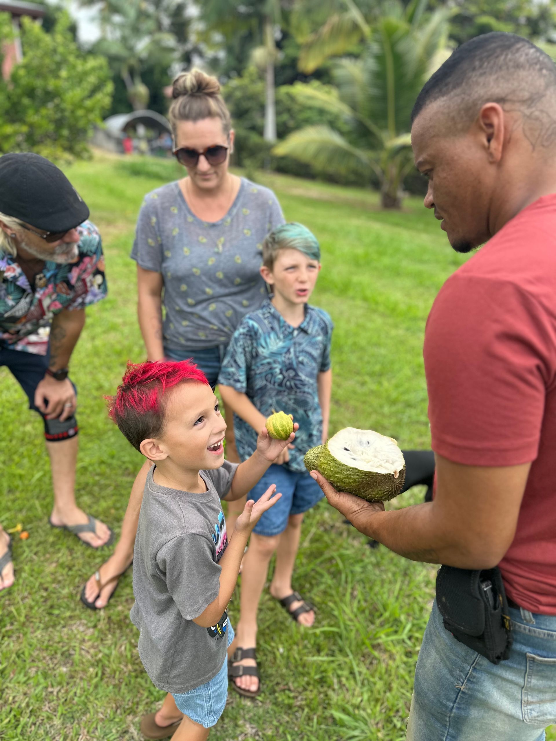 A man showing a custard apple to a group of people outdoors. A child with red hair is excited.