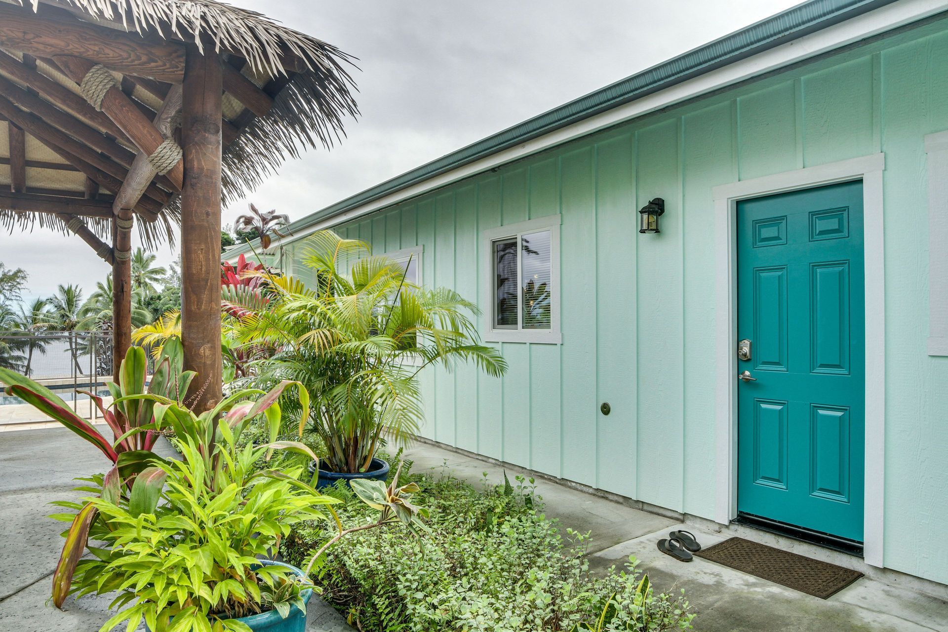 Exterior of a light blue building with a teal door, window, and tropical plants under a thatched roof structure.