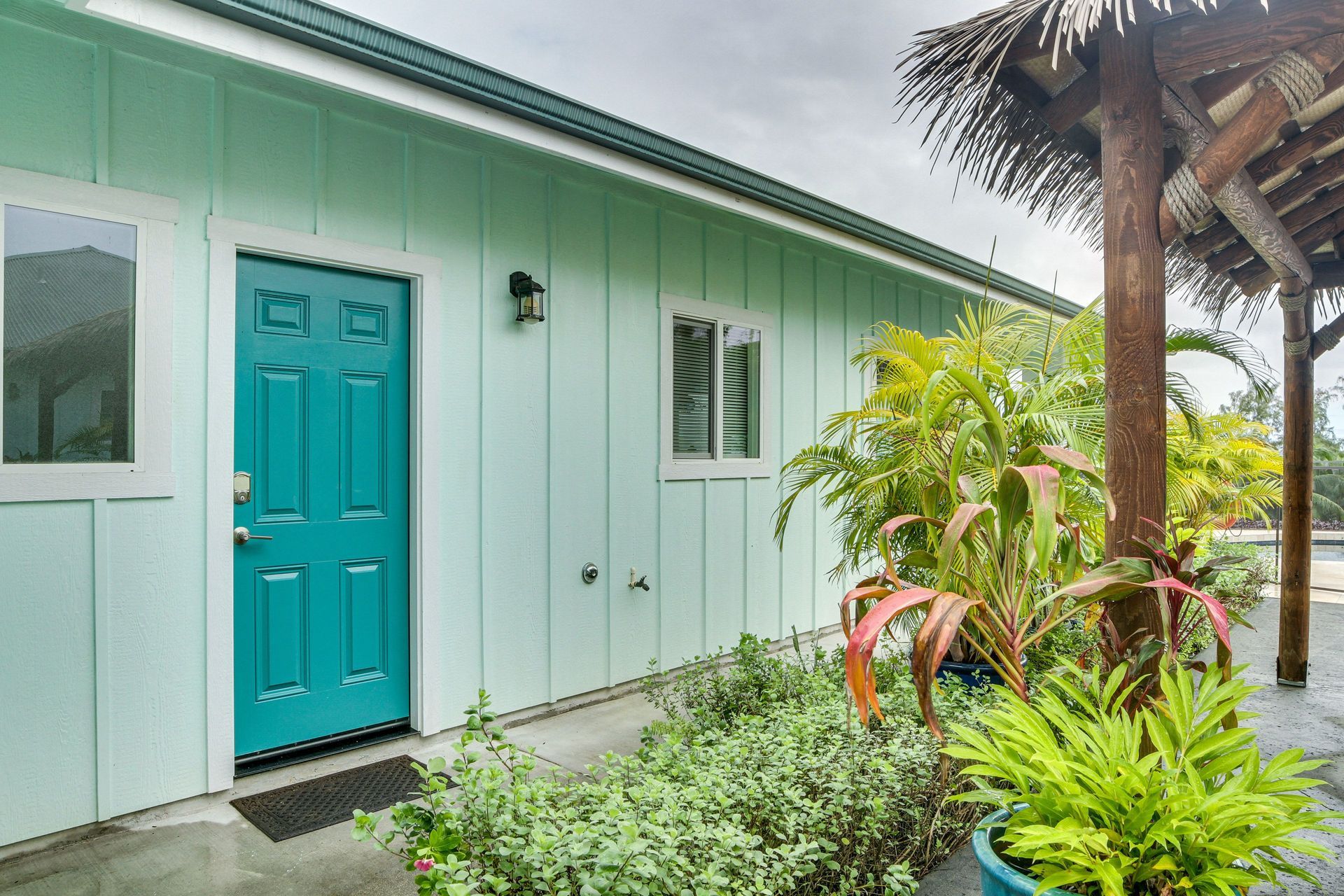 Turquoise door on a pale green building with tropical plants and a thatched roof pergola.