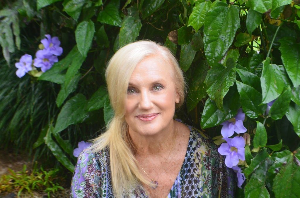 Blonde woman smiles, in front of leafy green plants with purple flowers.