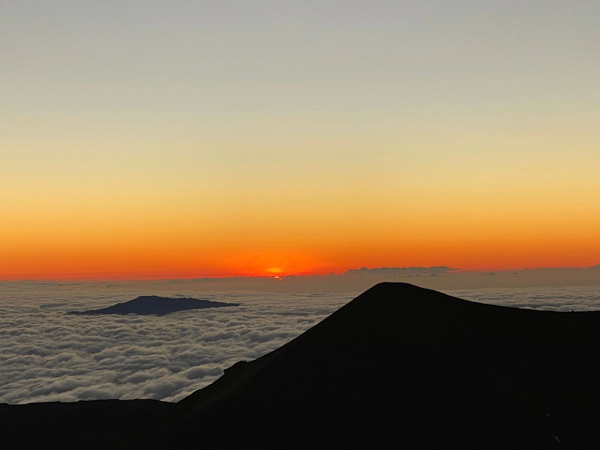 Sunrise over a sea of clouds, viewed from a mountain peak. Orange and gold hues dominate the sky.