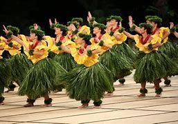 Dancers in yellow tops and green skirts perform hula. Stage with lines, dark backdrop.