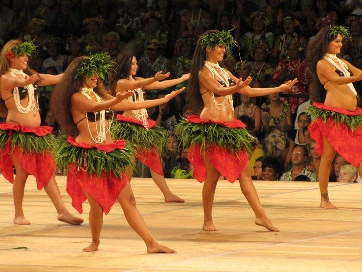 Hula dancers in red skirts and leaf garlands perform on a stage before an audience.