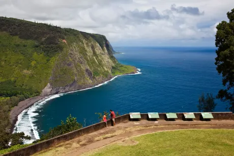 Coastal view of high cliffs and dark blue ocean; two people stand on a lookout point with grass.