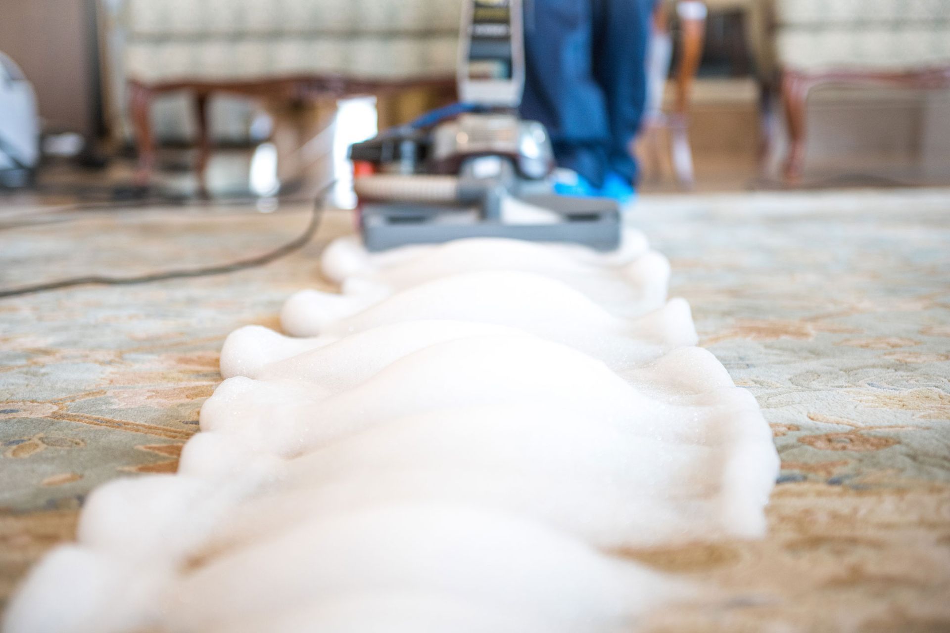 A cleaning machine spreads foam while moving across a carpet near some furniture. A cleaning machine spreads foam while moving across a carpet near some furniture.