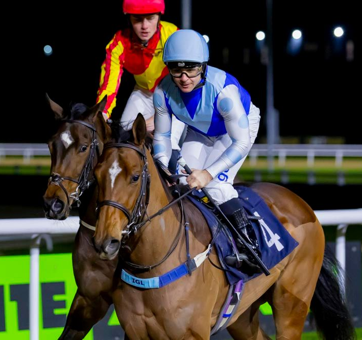 Two horses racing at night; jockeys in colorful silks.