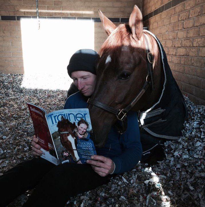 A man sitting next to a horse reading a trainer magazine