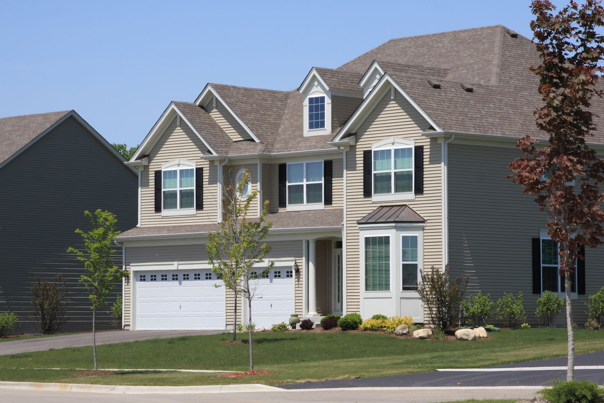 Two-story house with beige siding, white garage doors, black shutters, and a well-kept lawn under a blue sky.