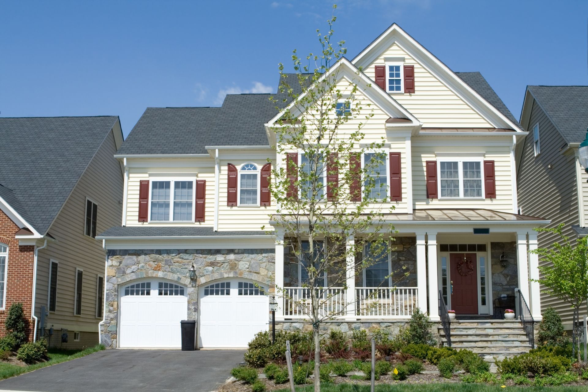 Two-story house with tan siding, stone facade, and red shutters, with a driveway and garage, in a neighborhood.