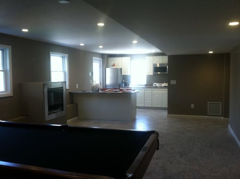 Empty basement rec room with gray walls, carpet, recessed lights, and a small kitchen area in back