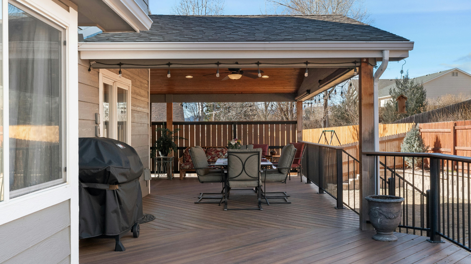 Covered backyard deck with grill, dining table, chairs, and railing overlooking a fenced yard.