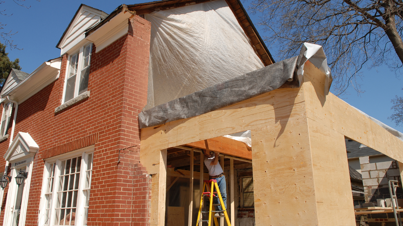 House under renovation with tan siding, brick walls, and a worker on a ladder by the porch