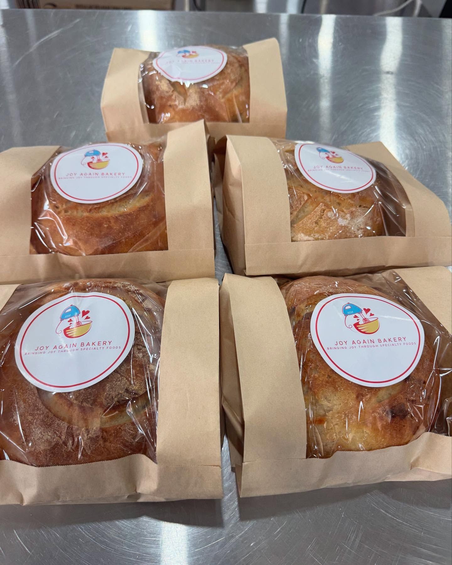 Five baked goods in brown paper bags, each with a clear window and a round label, on a metal surface.