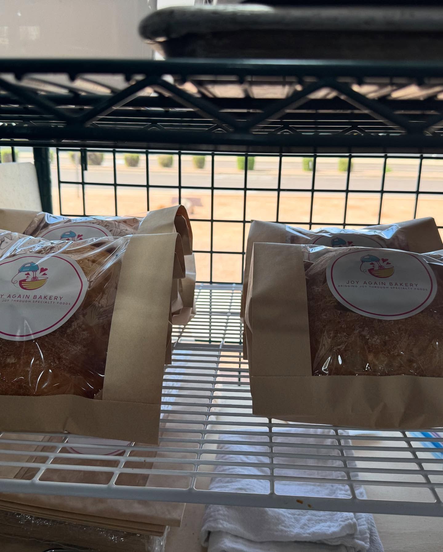 Bags of baked goods on a metal wire shelf, possibly in a bakery. Sunlight through a window.