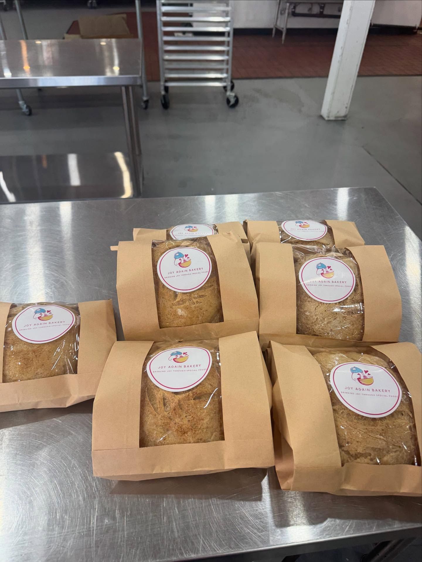 Loaves of bread in brown paper bags with clear window, stacked on a metal table.