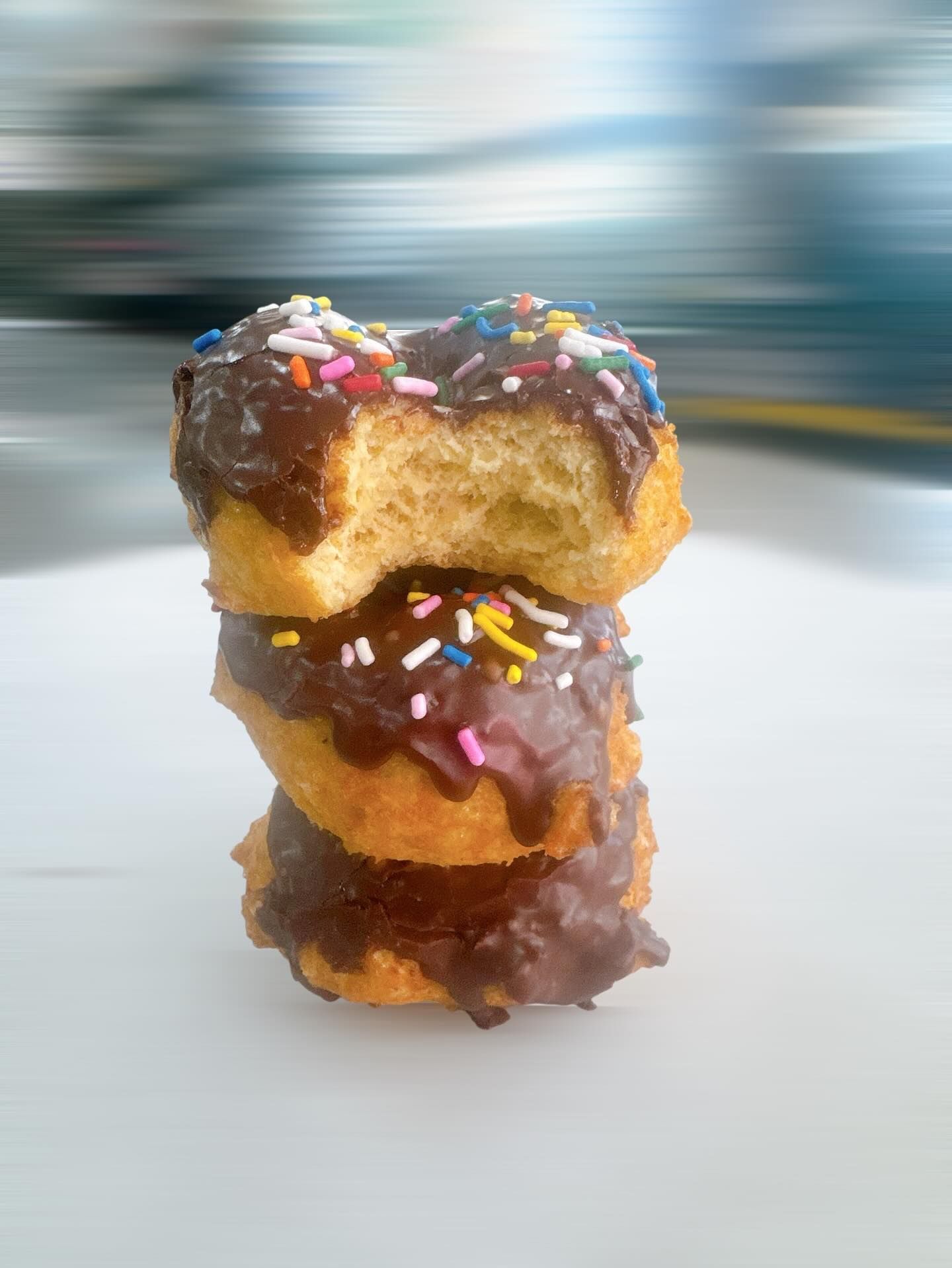 Stack of three chocolate-frosted donuts with sprinkles, one partially eaten, on a white surface.