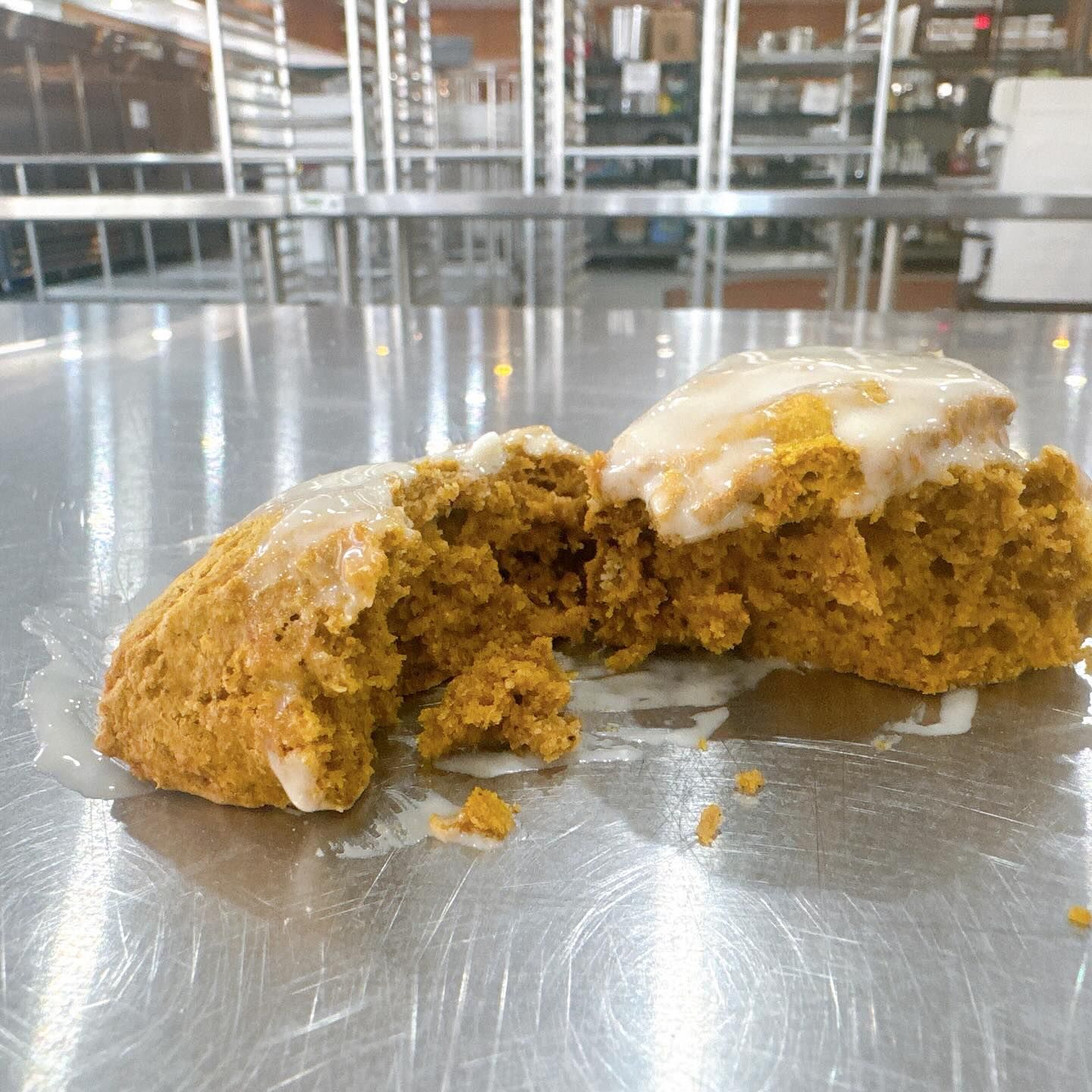 Pumpkin scone, cut in half, with icing, on a stainless steel surface. Blurred industrial kitchen in background.