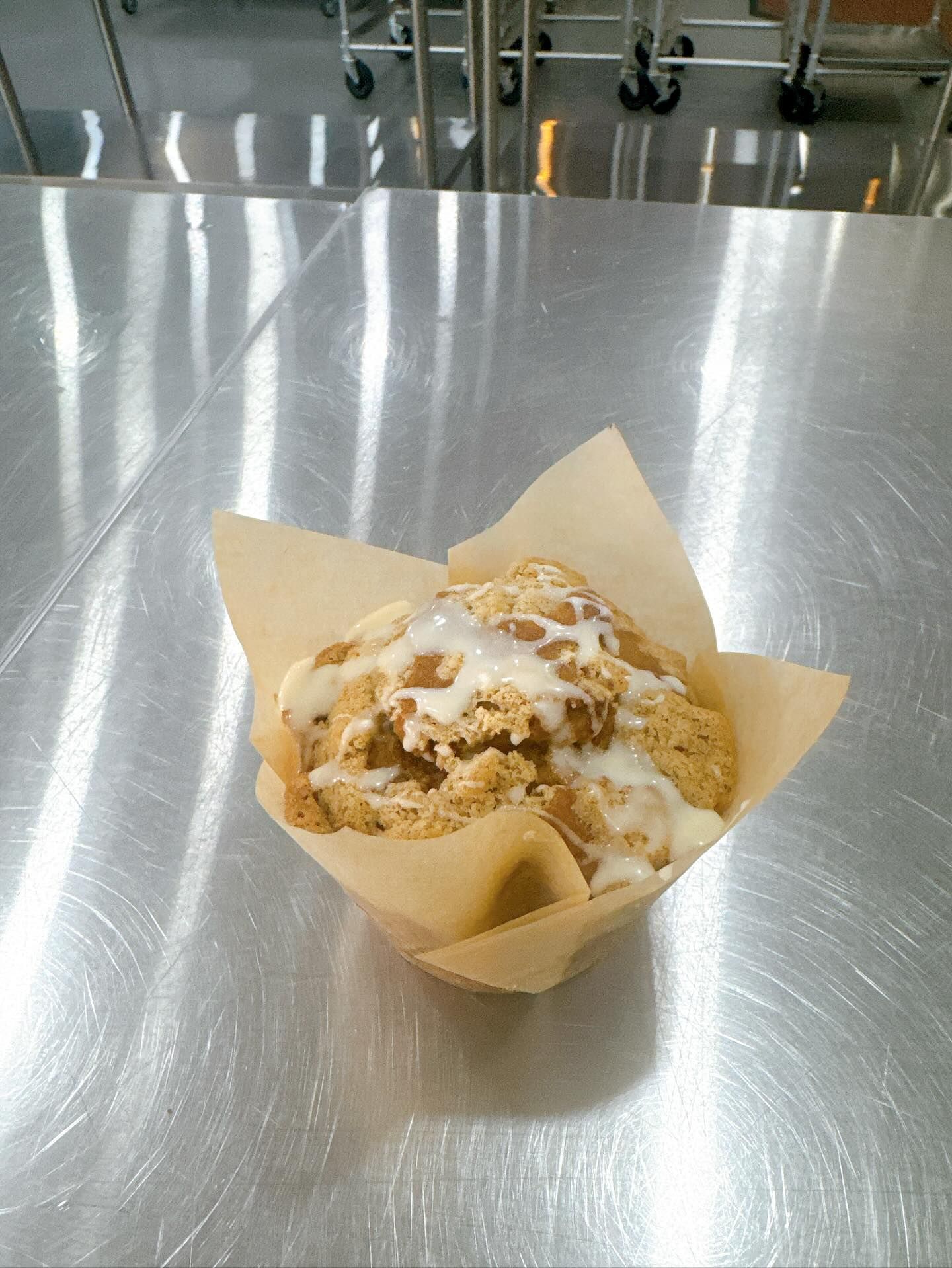 Muffin in brown paper holder, topped with icing and crumble, on a stainless steel surface.