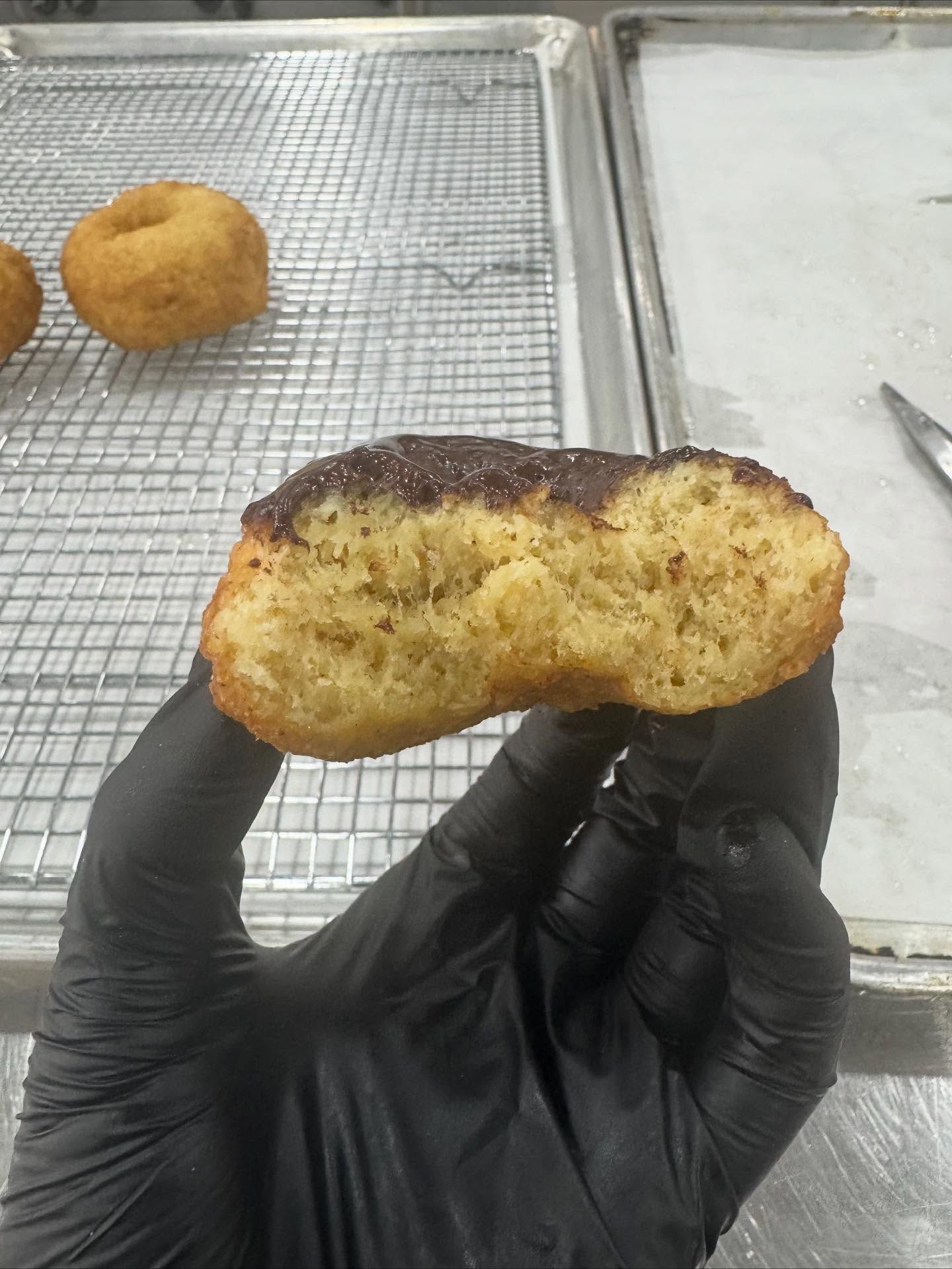 Hand holding a cut donut with a chocolate glaze, showing its soft interior, alongside other donuts on a cooling rack.