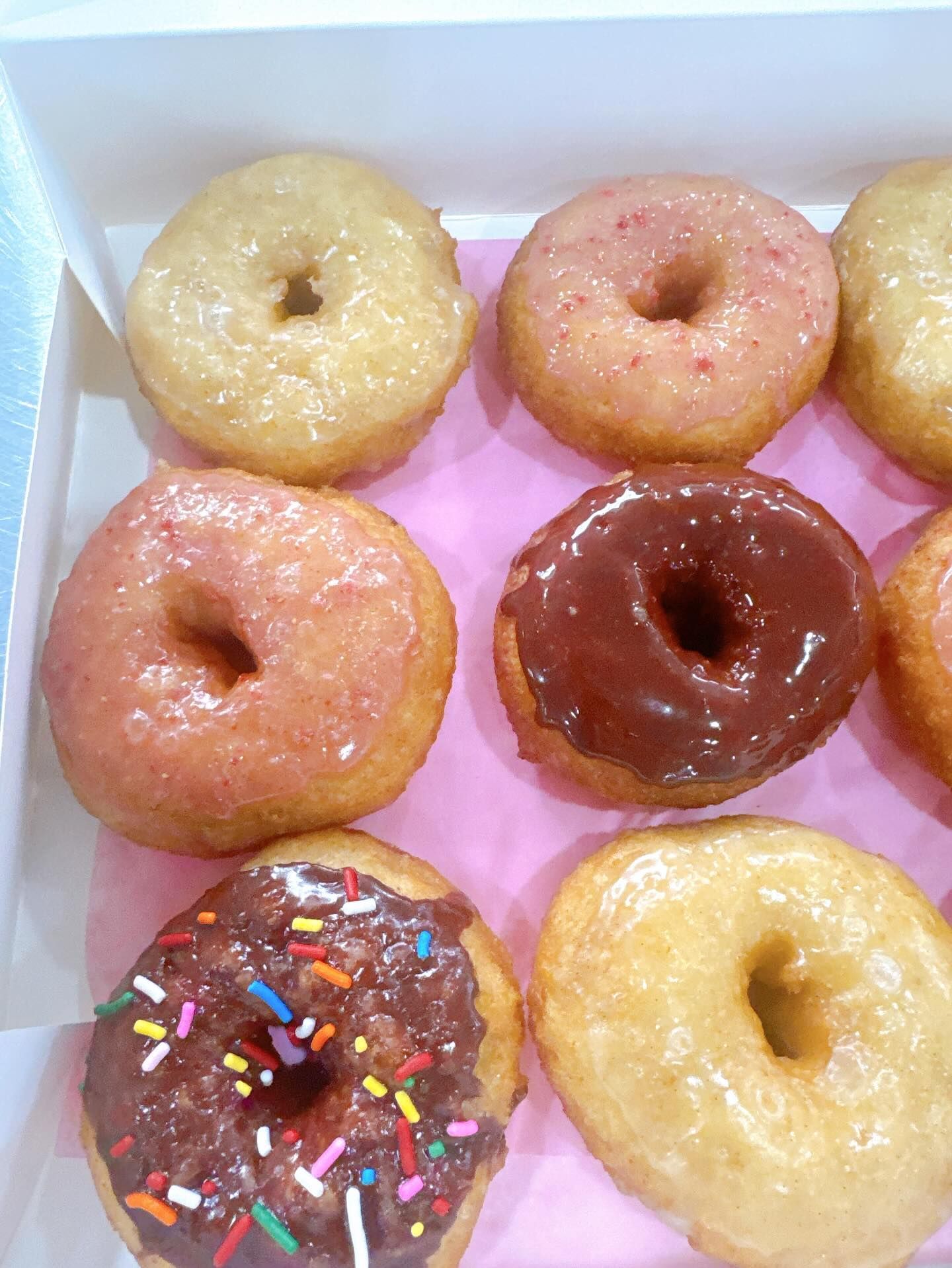Box of glazed donuts with various toppings: sprinkles, pink glaze, and chocolate.