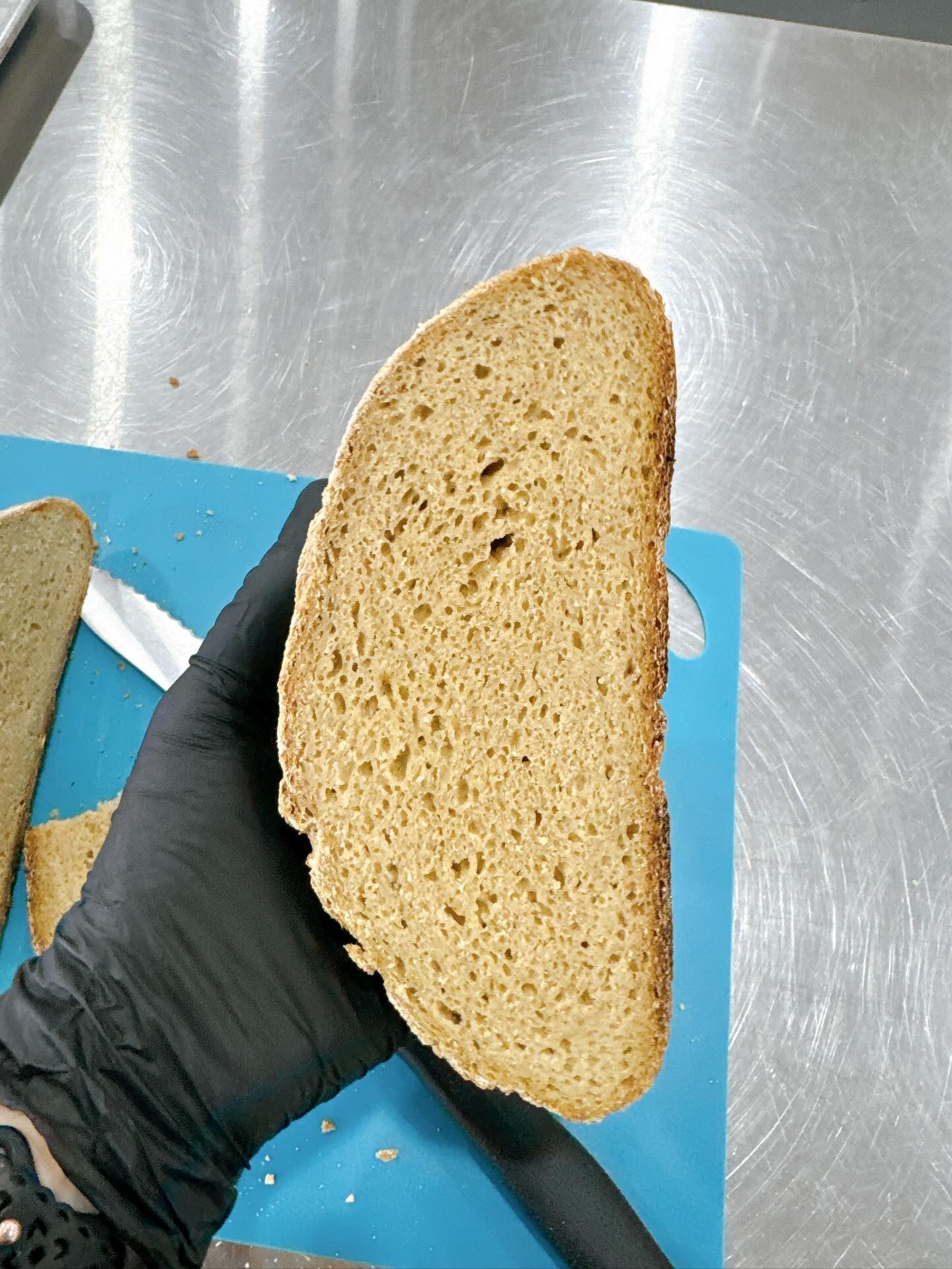 Hand holding a thick slice of brown bread; resting on a blue cutting board, stainless steel surface.