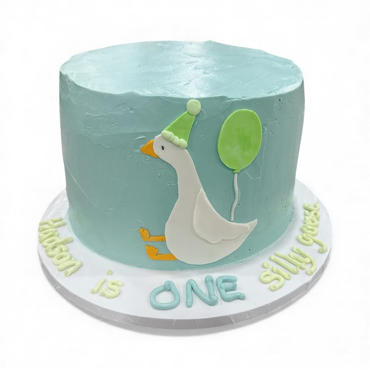 A teal frosted cake with a cartoon goose wearing a party hat, balloon, and