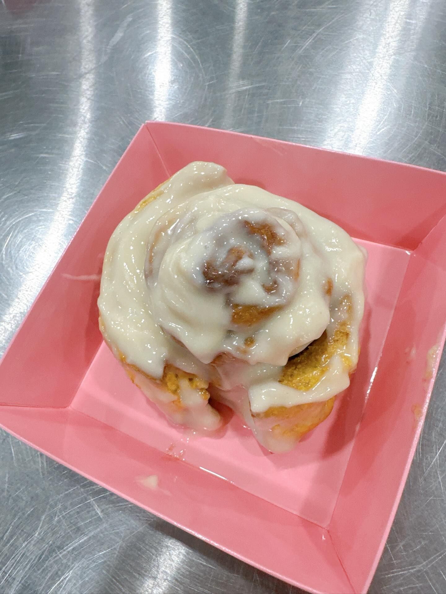 Cinnamon roll with white icing in a pink paper container on a metal surface.