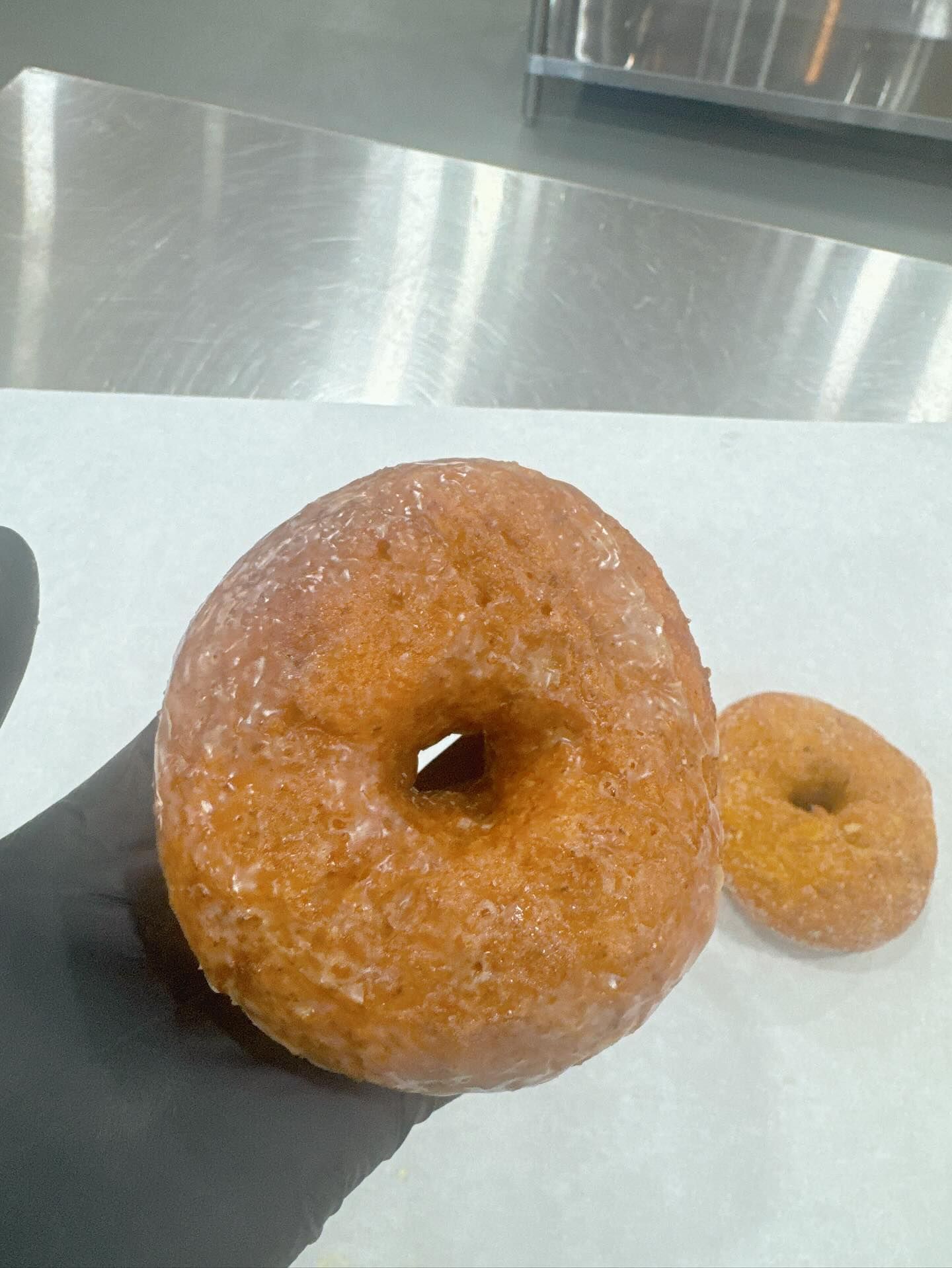 Glazed donut held in gloved hand, another donut in background on white paper.