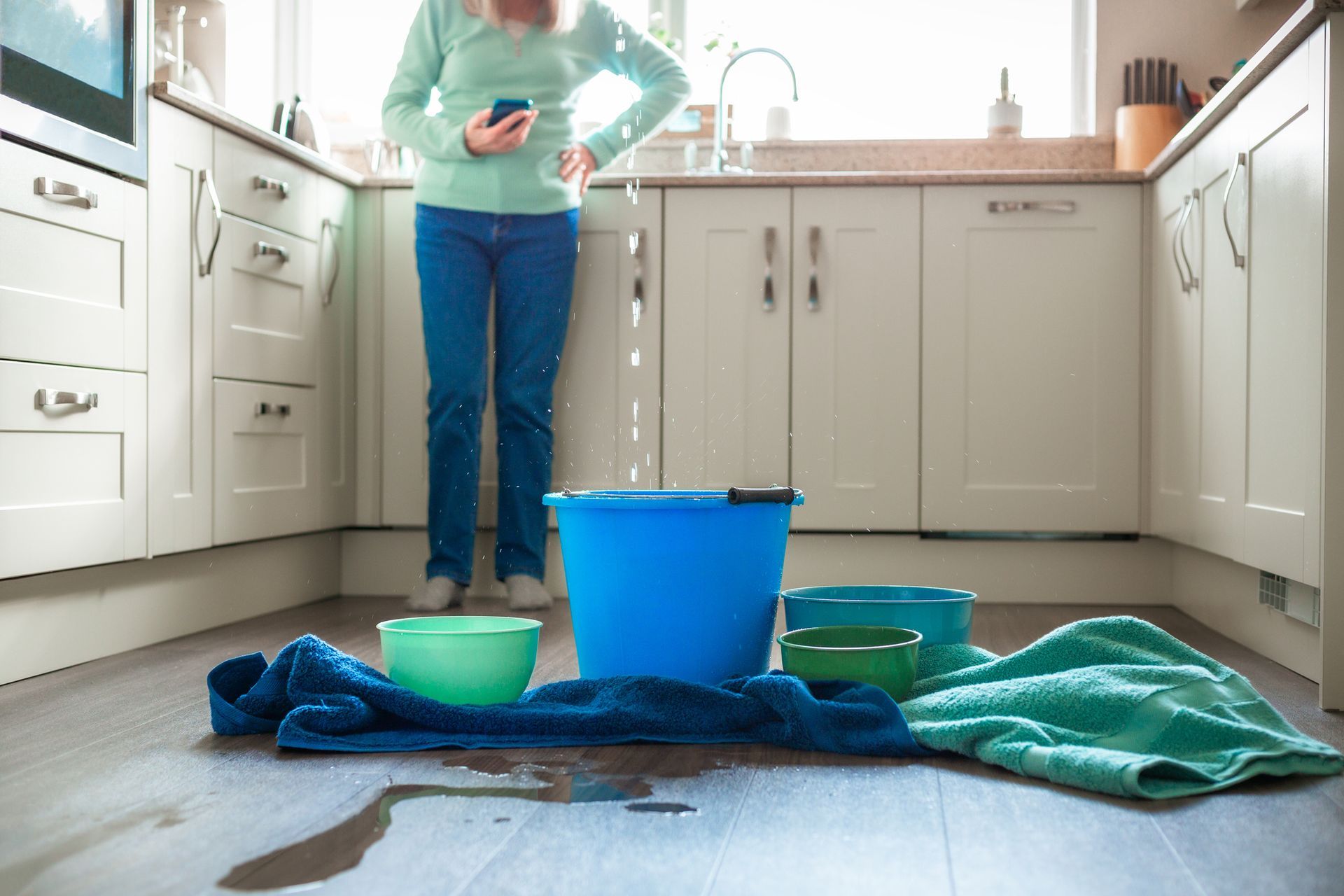 Woman in kitchen with water leaking from ceiling into buckets and towels on floor.