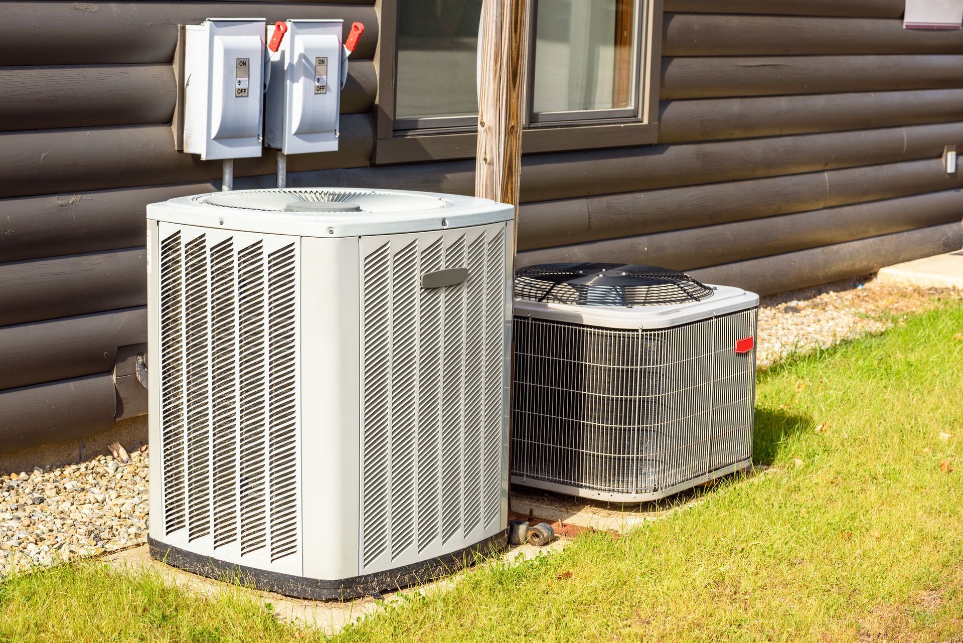 Two air conditioning units beside a wooden building, with electrical boxes on the wall and grass in front.
